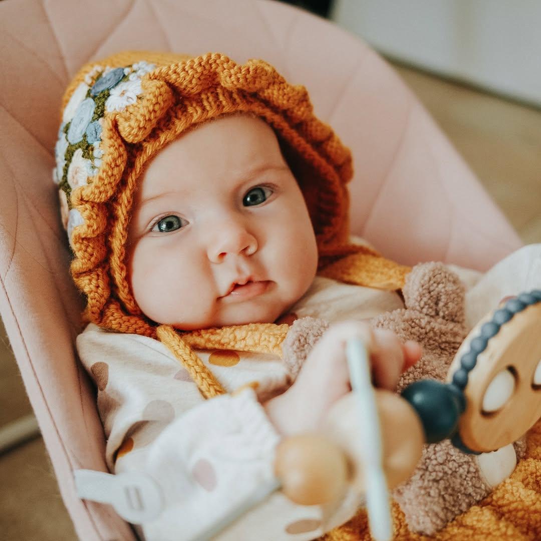 Baby in a polka dot onesie wearing a knitted orange bonnet with floral details, sitting in a pink chair, holding a toy and a plush bear.