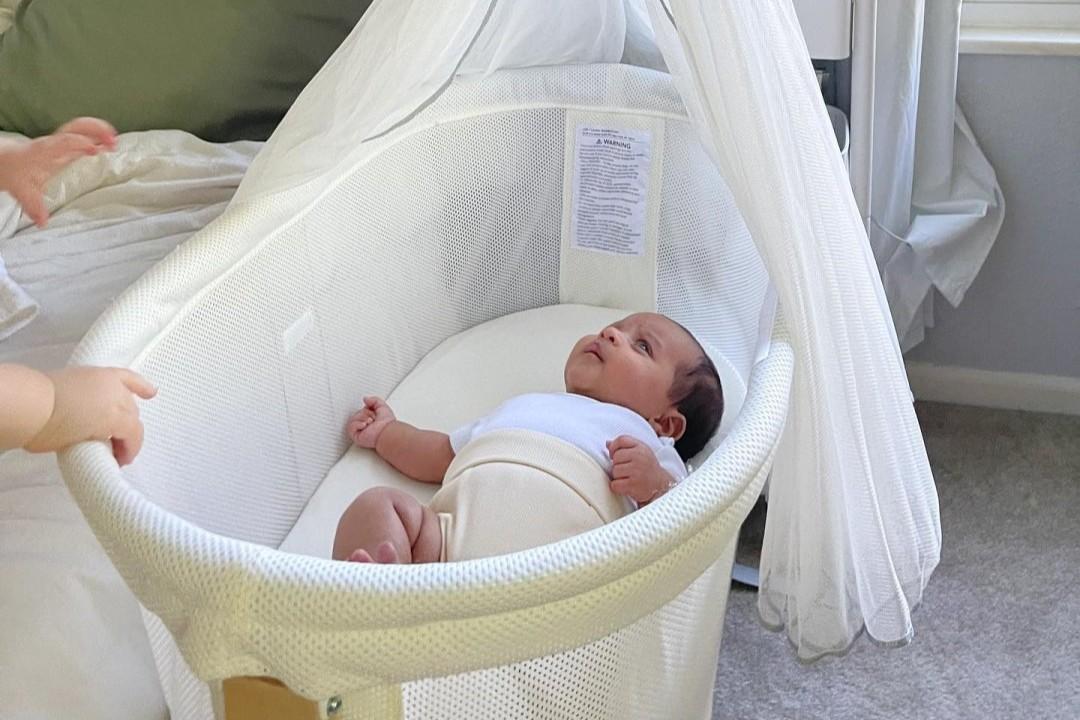 A baby lying in a white bassinet, looking up, with a sheer canopy overhead. The setting is a softly lit room with carpeted flooring.