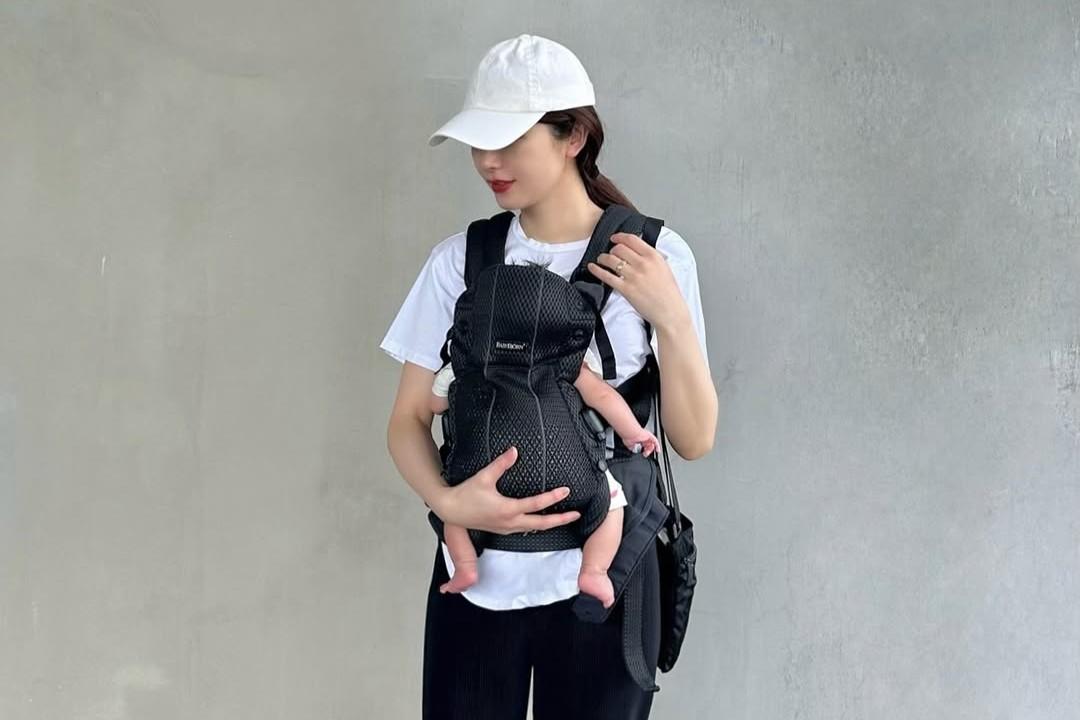 Woman wearing a cap and white t-shirt, carrying a baby in a black carrier against a plain gray background.