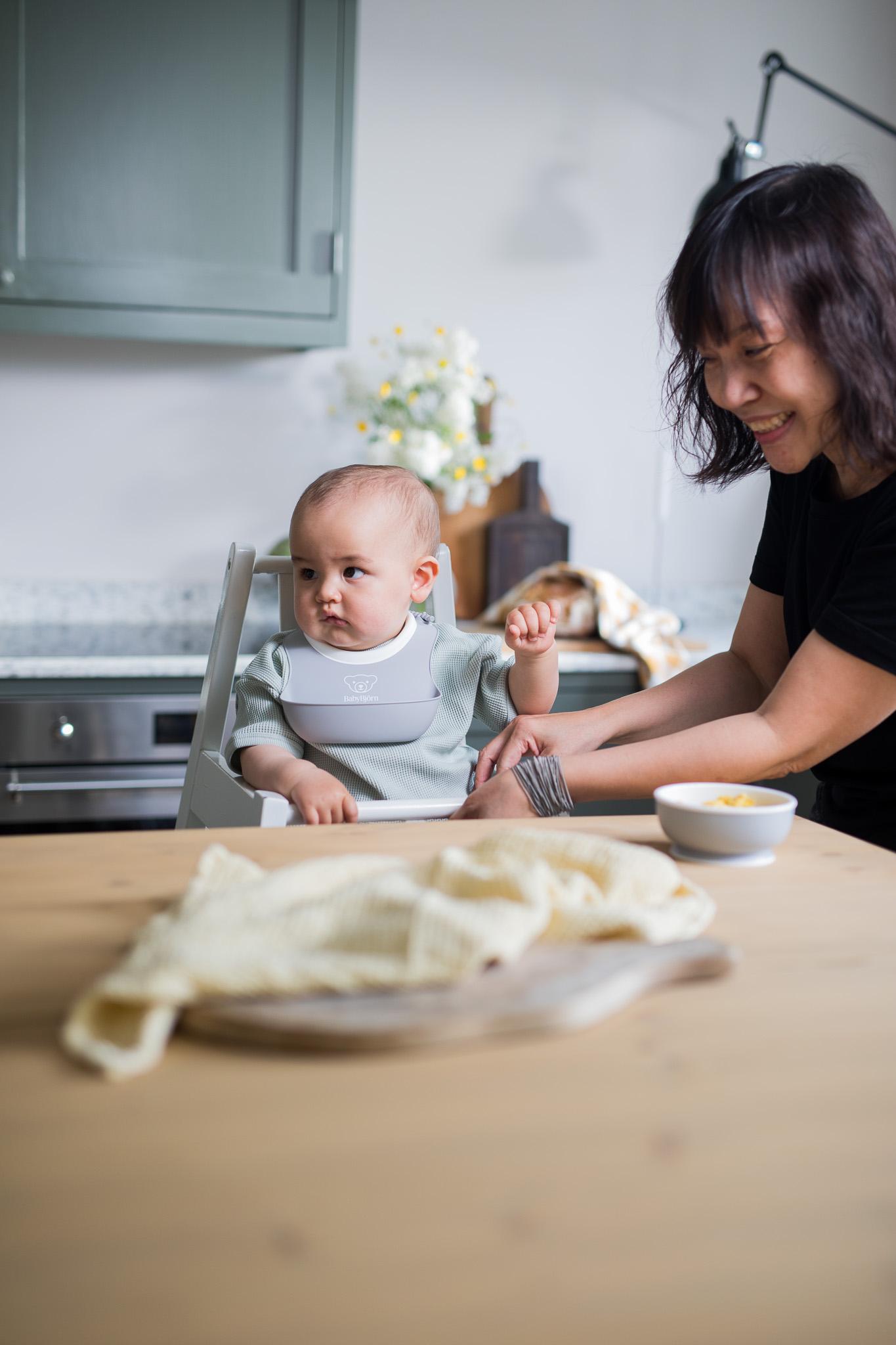 A smiling woman helps a baby wearing a bib in a high chair at a kitchen table with a bowl and cloth in the foreground.