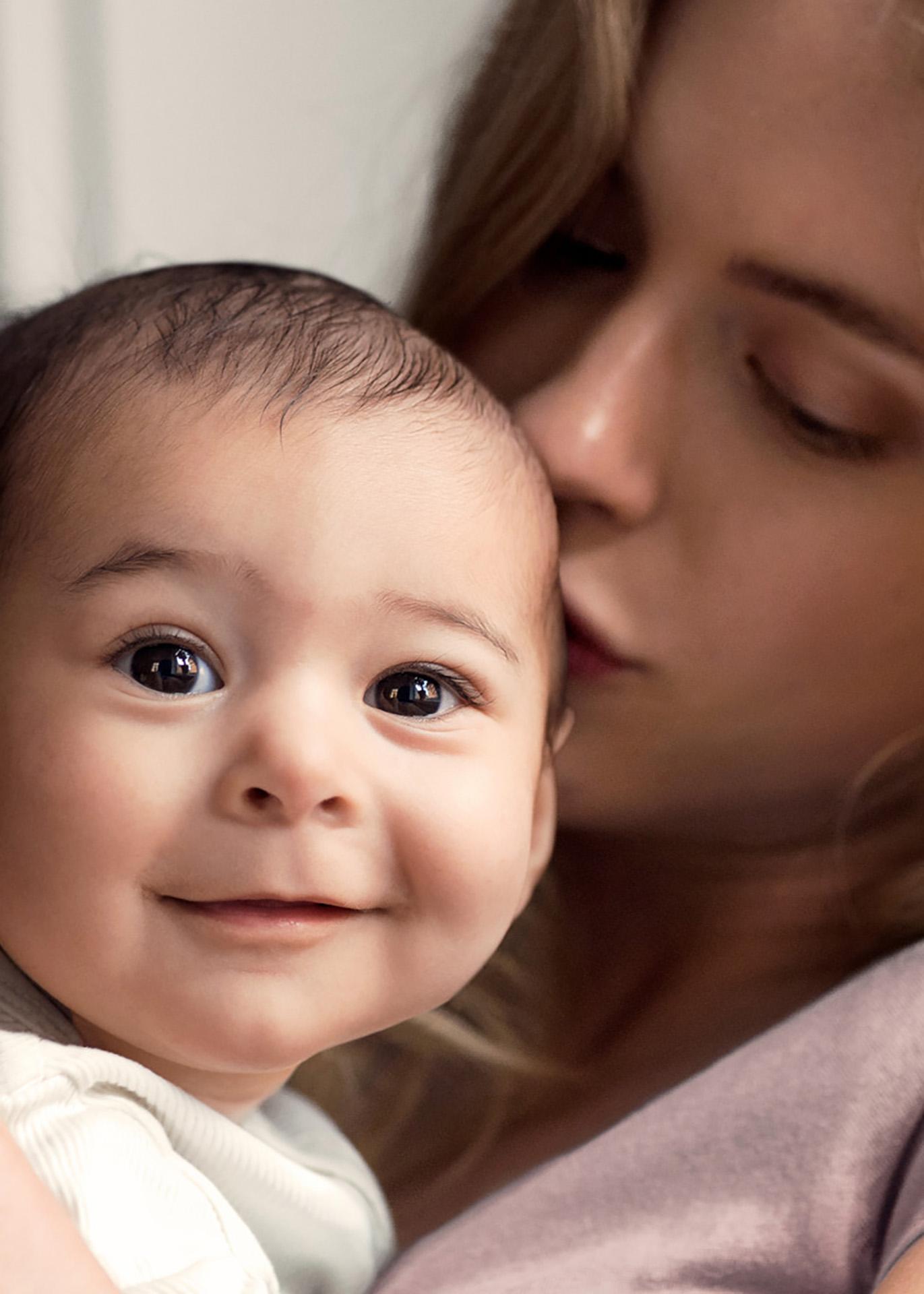 A smiling baby with big eyes is held close by a woman with long hair, expressing warmth and affection.