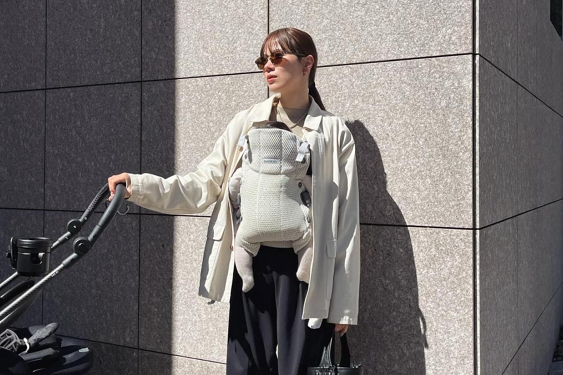 A person wearing sunglasses carries a baby in a front carrier while pushing a stroller, standing against a tiled wall in sunlight.