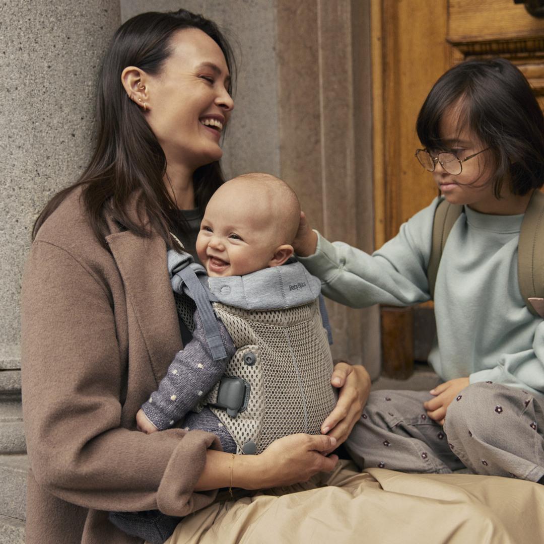 A smiling woman holds a baby in a carrier, while a child with glasses gently touches the baby's head. They are sitting near a stone column.