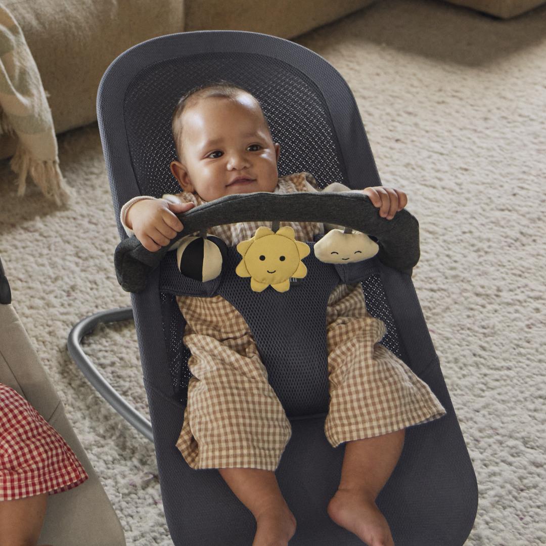 Baby in a checkered outfit sits in a bouncer holding a toy bar with plush sun and cloud, on a light carpeted floor.