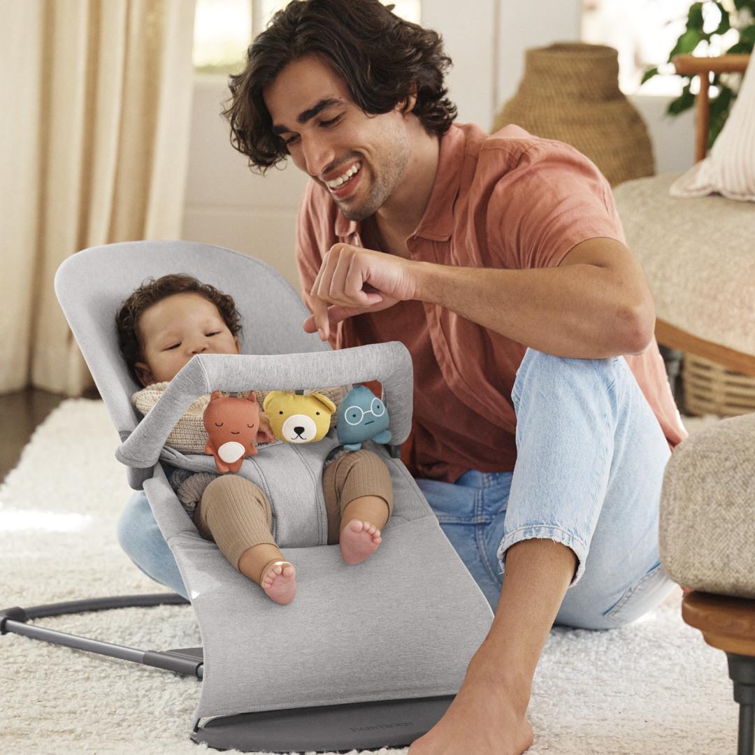 A smiling man interacts with a baby in a gray baby bouncer, surrounded by plush toys. The room is cozy with soft lighting.