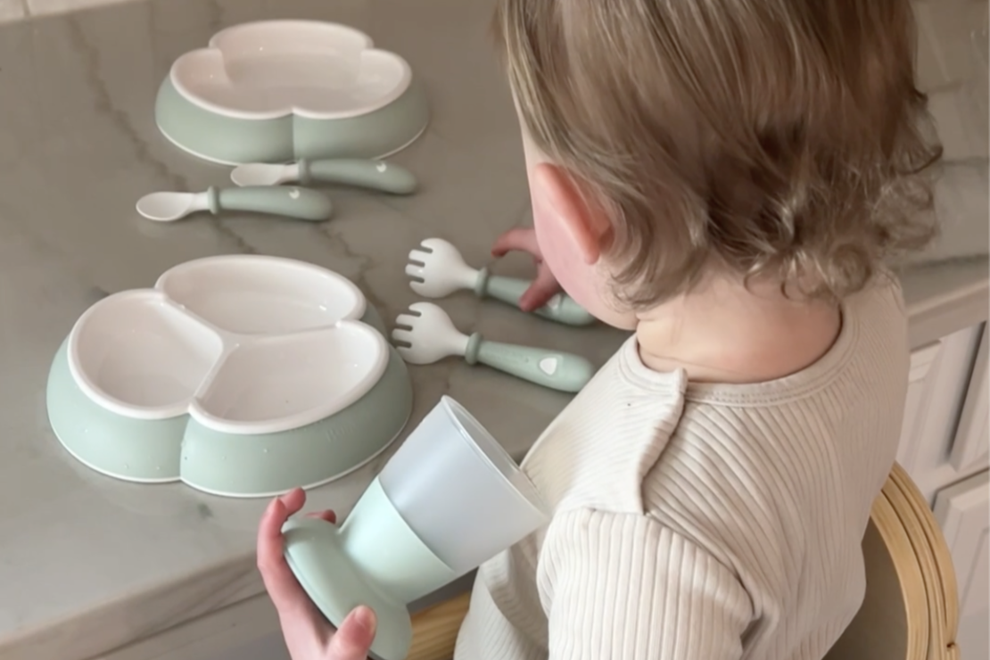 A toddler holds a green cup, sitting near a set of divided plates and utensils on a kitchen counter.