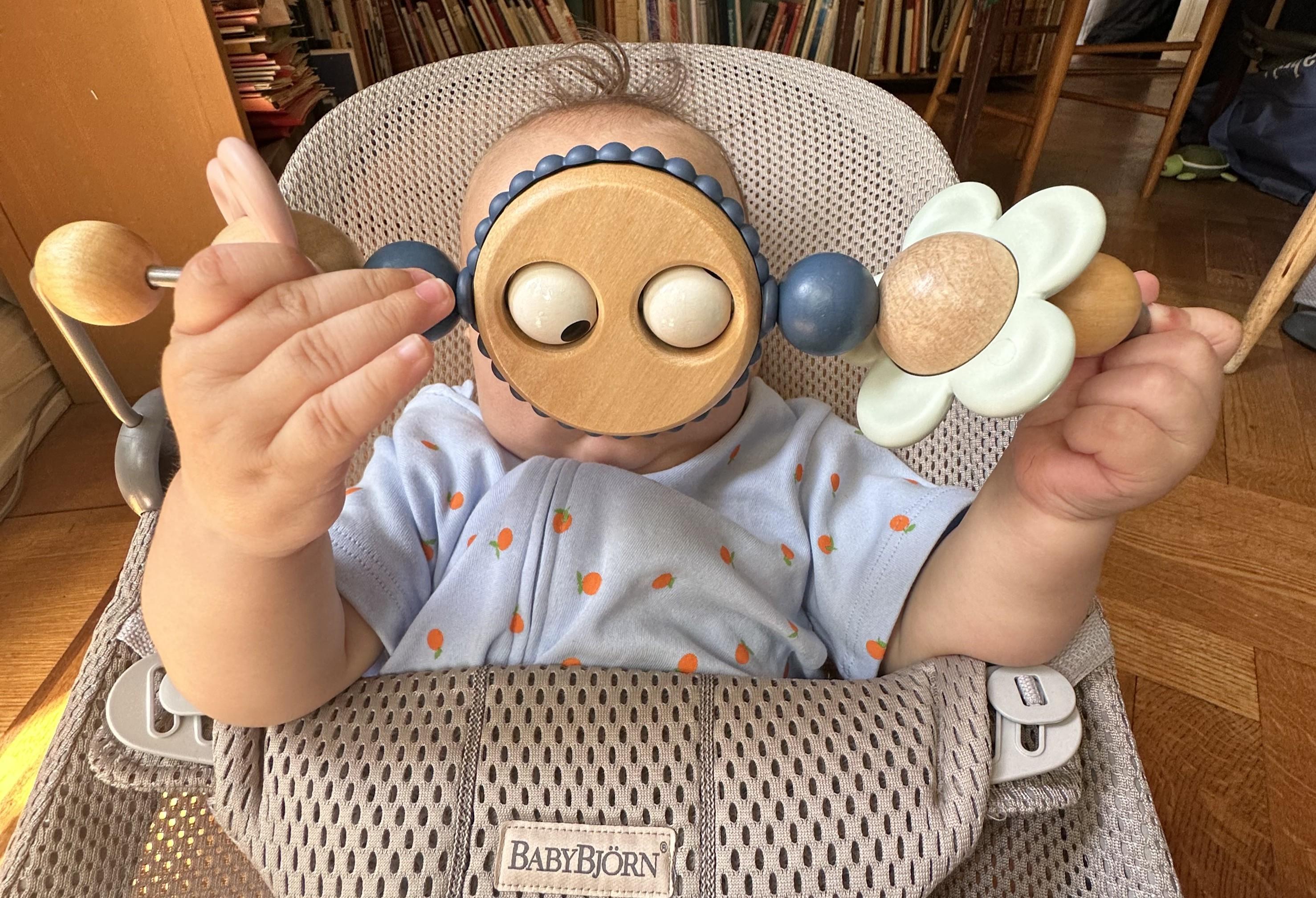 Baby in a high chair holding a wooden toy in front of their face, wearing a light blue outfit with orange dots.