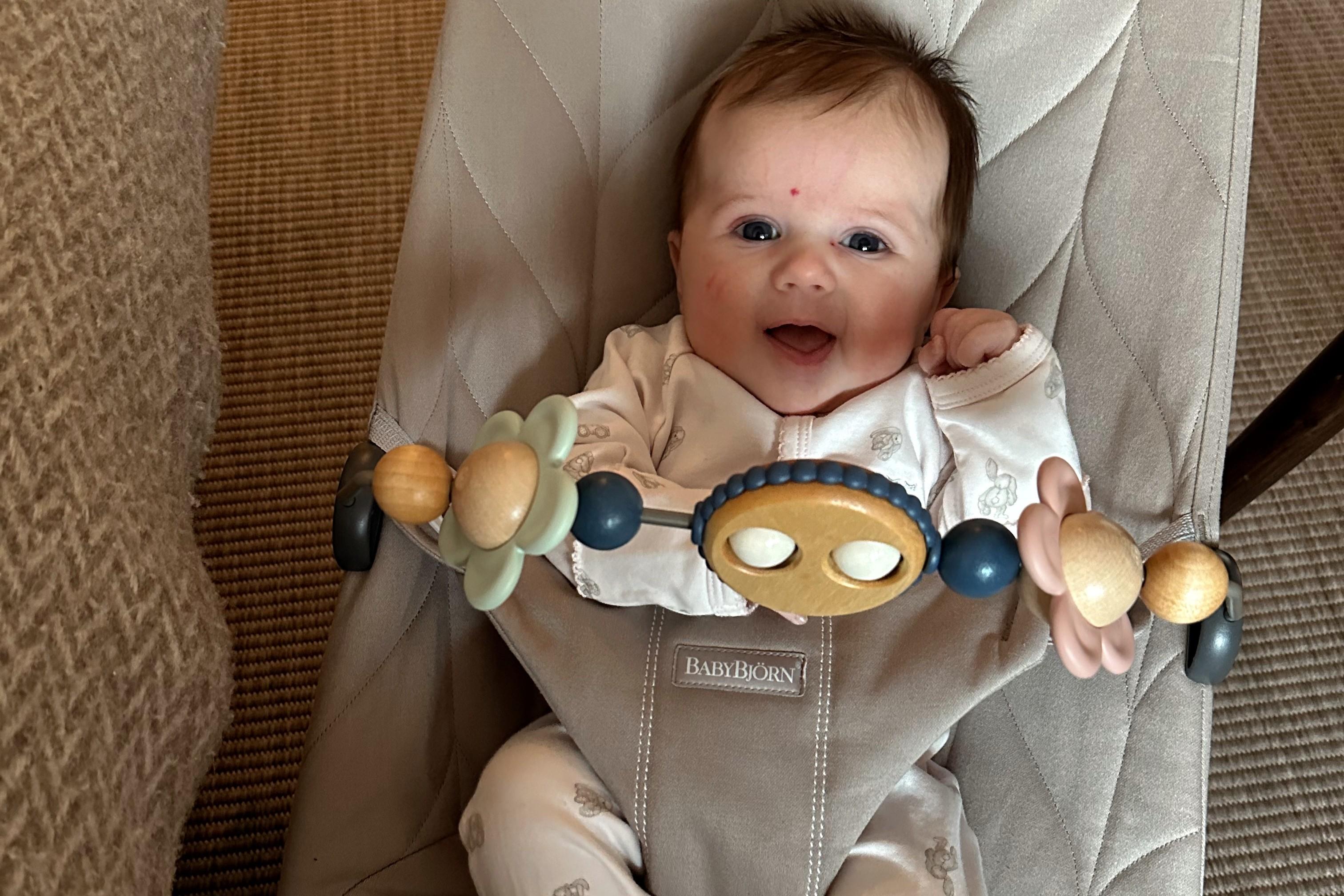 A smiling baby sits in a beige bouncer with a wooden and pastel-colored toy bar across the seat.