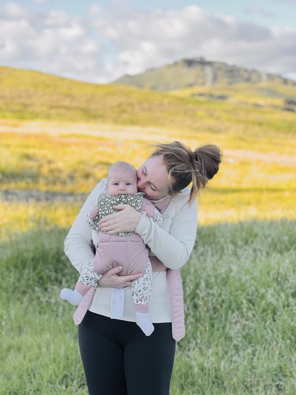 A person lovingly holds and kisses a baby, standing in a grassy field with hills and a cloudy sky in the background.